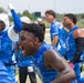 A group of athletes wearing blue camouflage uniforms labeled 'Air Force' are celebrating intensely on a football sideline. The central figure is shouting with an expression of intense emotion, while teammates behind him raise their arms in celebration. The setting appears to be an outdoor athletic field during a flag football or similar competition.