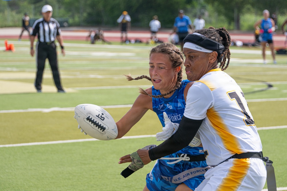 Two female athletes compete in a flag football game, with a U.S. Air Force player in a blue jersey attempting to pass or carry the ball while being defended by an opponent in a white and gold uniform. A referee in a black and white striped shirt is visible in the background, along with other players and spectators on what appears to be an outdoor turf field. The action is captured in sharp focus with a shallow depth of field.