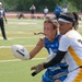 Two female athletes compete in a flag football game, with a U.S. Air Force player in a blue jersey attempting to pass or carry the ball while being defended by an opponent in a white and gold uniform. A referee in a black and white striped shirt is visible in the background, along with other players and spectators on what appears to be an outdoor turf field. The action is captured in sharp focus with a shallow depth of field.