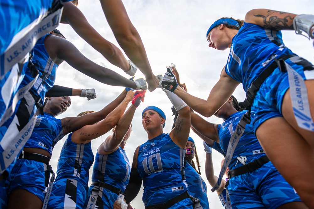 A U.S. Air Force women's flag football team huddles together in a pre-game or motivational team circle, raising their hands to the center in a show of unity. The players are wearing blue Air Force branded uniforms with flag football belts. The photo is taken from a low angle looking upward, creating a dynamic and dramatic perspective.