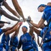 A U.S. Air Force women's flag football team huddles together in a pre-game or motivational team circle, raising their hands to the center in a show of unity. The players are wearing blue Air Force branded uniforms with flag football belts. The photo is taken from a low angle looking upward, creating a dynamic and dramatic perspective.