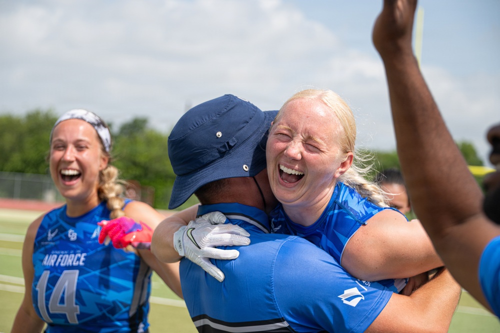 A celebratory moment on a sports field featuring players wearing U.S. Air Force uniforms in blue. A blonde woman is joyfully embracing a person wearing a navy bucket hat and white glove, both laughing. In the background, another player wearing jersey number 14 is also laughing and celebrating.