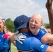 A celebratory moment on a sports field featuring players wearing U.S. Air Force uniforms in blue. A blonde woman is joyfully embracing a person wearing a navy bucket hat and white glove, both laughing. In the background, another player wearing jersey number 14 is also laughing and celebrating.