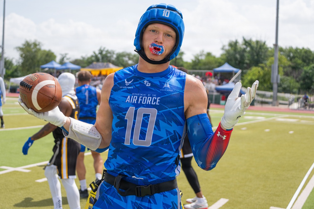 A male athlete wearing a blue 'Department of the Air Force' jersey with number 10 poses on a football field holding a football in one hand while gesturing with the other. He is equipped with a blue padded helmet, white gloves, a red/blue mouthguard, and a black belt, indicating this is a flag football game. The background shows other players, blue canopy tents, spectators, and a well-maintained athletic facility.