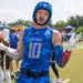 A male athlete wearing a blue 'Department of the Air Force' jersey with number 10 poses on a football field holding a football in one hand while gesturing with the other. He is equipped with a blue padded helmet, white gloves, a red/blue mouthguard, and a black belt, indicating this is a flag football game. The background shows other players, blue canopy tents, spectators, and a well-maintained athletic facility.