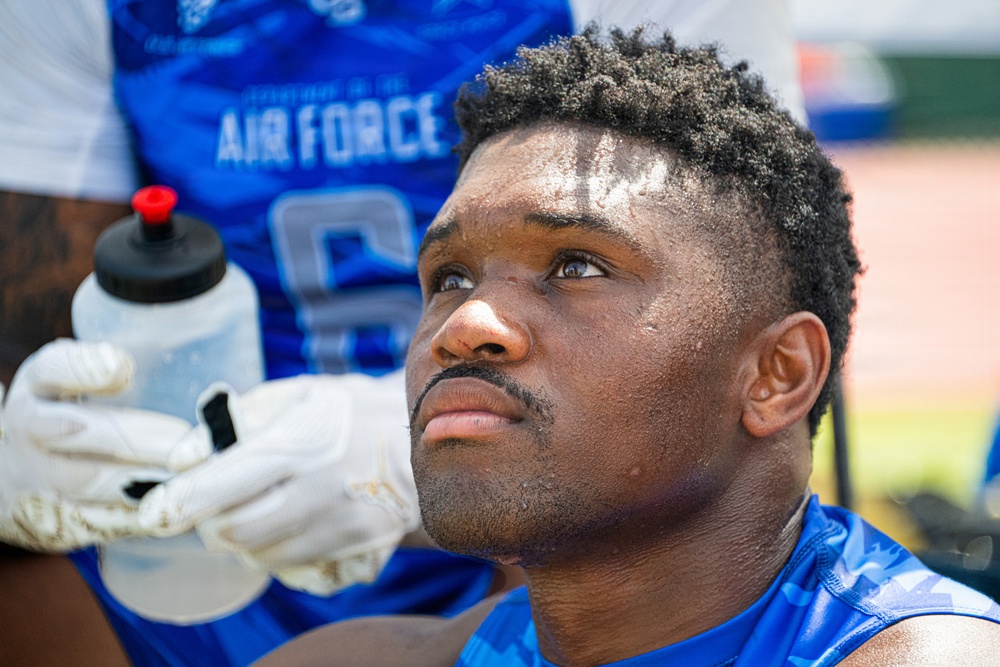 A close-up photograph of a male athlete wearing a blue Air Force football jersey, appearing fatigued or contemplative during what seems to be a game or practice session. His face shows visible sweat and exertion, and he is looking upward. A gloved hand holding a water bottle is visible in the foreground.