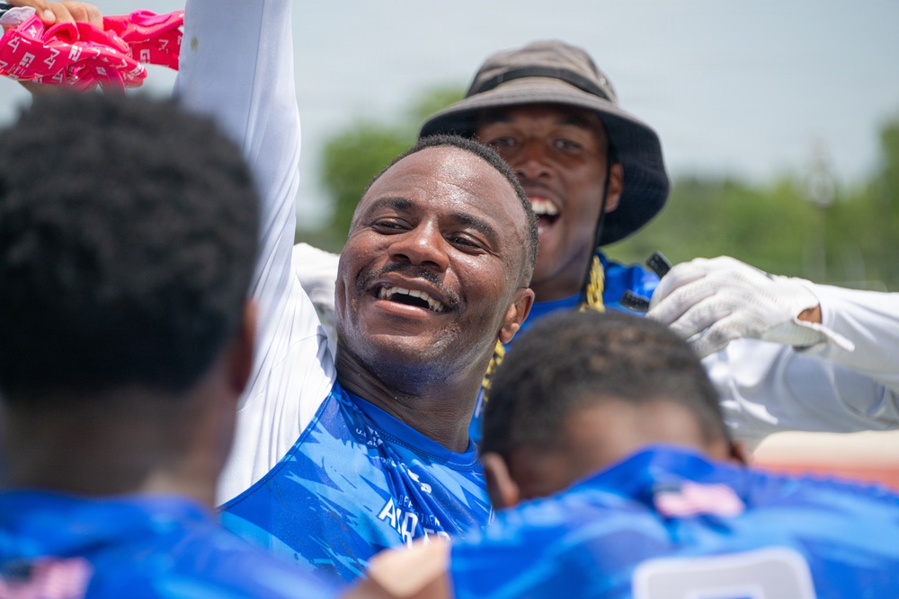 A group of individuals wearing blue athletic jerseys are gathered together in what appears to be a celebratory huddle or team moment. The central figure is smiling broadly with one arm raised, expressing joy or excitement. The setting appears to be an outdoor sports event on a sunny day.