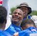 A group of individuals wearing blue athletic jerseys are gathered together in what appears to be a celebratory huddle or team moment. The central figure is smiling broadly with one arm raised, expressing joy or excitement. The setting appears to be an outdoor sports event on a sunny day.