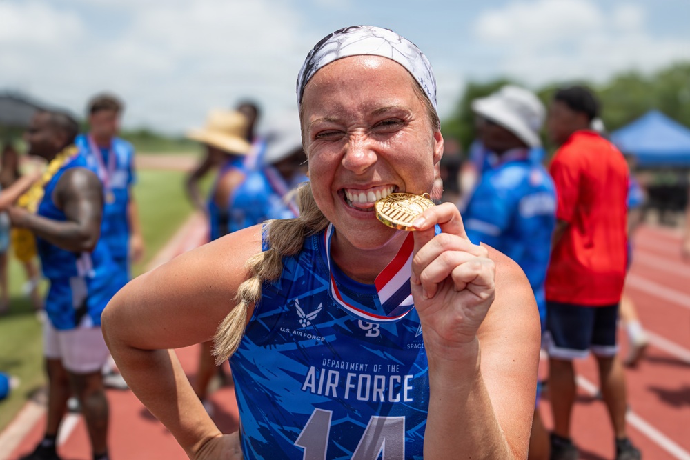 A woman wearing a U.S. Air Force Department of the Air Force jersey (number 14) joyfully bites a gold medal on what appears to be an outdoor athletic track. She is celebrating a victory at what looks like a military sports competition, with other athletes in blue jerseys visible in the blurred background. The image captures a classic celebratory pose commonly seen at sporting events.