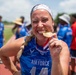 A woman wearing a U.S. Air Force Department of the Air Force jersey (number 14) joyfully bites a gold medal on what appears to be an outdoor athletic track. She is celebrating a victory at what looks like a military sports competition, with other athletes in blue jerseys visible in the blurred background. The image captures a classic celebratory pose commonly seen at sporting events.