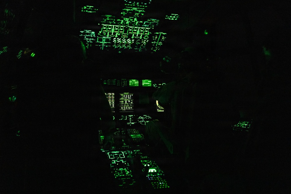 The image shows an aircraft cockpit photographed under night vision compatible green lighting conditions. Multiple instrument panels, overhead panels, and display screens are visible, illuminated in green to be compatible with night vision goggles (NVGs). At least two crew members are silhouetted in the cockpit seats, suggesting active flight operations at night.