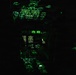 The image shows an aircraft cockpit photographed under night vision compatible green lighting conditions. Multiple instrument panels, overhead panels, and display screens are visible, illuminated in green to be compatible with night vision goggles (NVGs). At least two crew members are silhouetted in the cockpit seats, suggesting active flight operations at night.