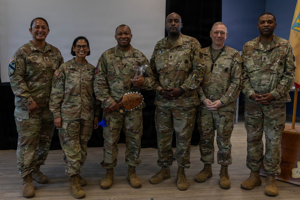 Six U.S. Army soldiers in OCP (Operational Camouflage Pattern) uniforms pose together for a group photo in what appears to be an indoor military facility. The central figure is holding what appears to be a decorative football trophy or award, suggesting this is an award or recognition ceremony. Name tapes visible on uniforms include 'BAILEY,' 'MALAVE,' and 'LEDET.'