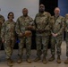 Six U.S. Army soldiers in OCP (Operational Camouflage Pattern) uniforms pose together for a group photo in what appears to be an indoor military facility. The central figure is holding what appears to be a decorative football trophy or award, suggesting this is an award or recognition ceremony. Name tapes visible on uniforms include 'BAILEY,' 'MALAVE,' and 'LEDET.'