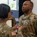 A U.S. Army promotion or award ceremony is taking place indoors. A female soldier is pinning a rank insignia or device onto the uniform of a male soldier identified by his name tape as 'Bailey.' A third soldier is visible in the background holding what appears to be an official certificate or order.