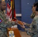Two U.S. Army soldiers in OCP (Operational Camouflage Pattern) uniforms are engaged in a presentation or award ceremony, with one soldier handing a decorative wooden item to the other. The setting appears to be an official military ceremony room with U.S. and other flags visible in the background. The male soldier on the left wears an 82nd Airborne Division patch, while the female soldier on the right wears a patch consistent with the 21st Theater Sustainment Command.