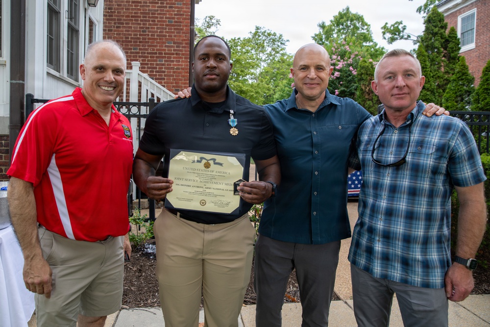 Four men pose together outdoors at what appears to be a military recognition ceremony. The second man from the left is holding a framed certificate reading 'Joint Service Achievement Medal' from the United States of America, and is wearing a medal on his chest. The setting appears to be in front of a brick building with greenery visible in the background.