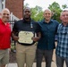 Four men pose together outdoors at what appears to be a military recognition ceremony. The second man from the left is holding a framed certificate reading 'Joint Service Achievement Medal' from the United States of America, and is wearing a medal on his chest. The setting appears to be in front of a brick building with greenery visible in the background.