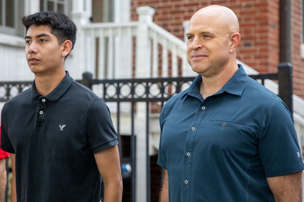 Two males are standing outdoors in front of a brick building with white stairs and a black iron fence. The younger male on the left wears a black American Eagle polo shirt, while the older bald male on the right wears a dark teal button-up shirt. Both appear to be looking in the same direction with neutral to serious expressions.