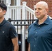 Two males are standing outdoors in front of a brick building with white stairs and a black iron fence. The younger male on the left wears a black American Eagle polo shirt, while the older bald male on the right wears a dark teal button-up shirt. Both appear to be looking in the same direction with neutral to serious expressions.