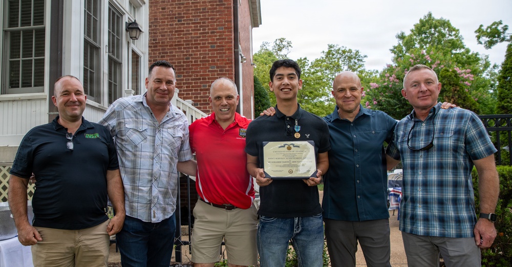 Six men are posing together outdoors in front of a brick building, with one younger man holding a framed certificate or award. The certificate appears to be an official U.S. government document, and the recipient is also wearing a medal around his neck. The group appears to be celebrating the young man's achievement at what looks like a casual outdoor gathering.