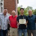 Six men are posing together outdoors in front of a brick building, with one younger man holding a framed certificate or award. The certificate appears to be an official U.S. government document, and the recipient is also wearing a medal around his neck. The group appears to be celebrating the young man's achievement at what looks like a casual outdoor gathering.
