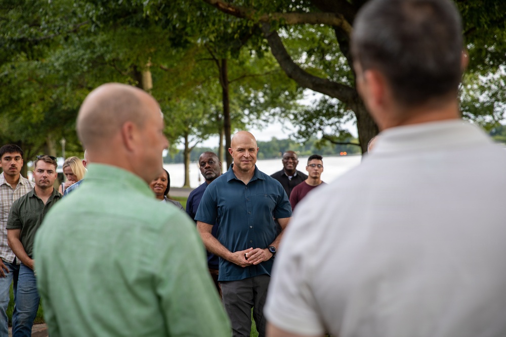 A group of people are gathered outdoors in a park-like setting near a body of water, with large trees providing shade. A man in a teal/dark blue short-sleeve button-up shirt stands centrally in focus, appearing to be the focal point of the gathering. The scene suggests an outdoor meeting, briefing, or community event.