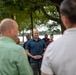 A group of people are gathered outdoors in a park-like setting near a body of water, with large trees providing shade. A man in a teal/dark blue short-sleeve button-up shirt stands centrally in focus, appearing to be the focal point of the gathering. The scene suggests an outdoor meeting, briefing, or community event.