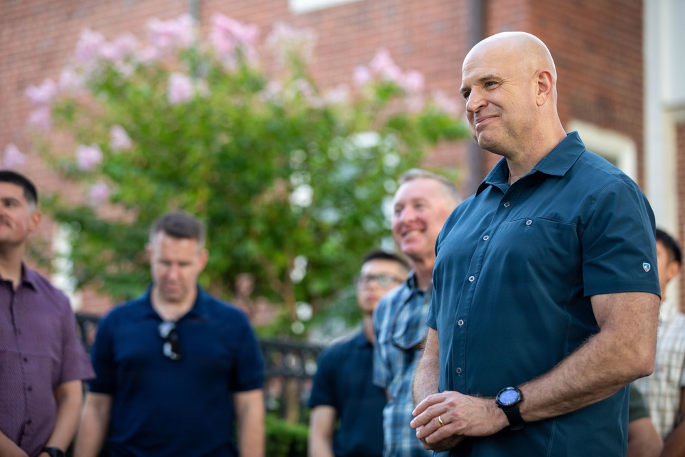 A bald man in a teal/dark blue short-sleeve button-up shirt stands in the foreground, smiling slightly, at what appears to be an outdoor gathering. Several other men are visible in the background, also smiling and appearing engaged in a social event. The setting includes a brick building and a flowering tree with pink blossoms.