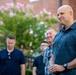 A bald man in a teal/dark blue short-sleeve button-up shirt stands in the foreground, smiling slightly, at what appears to be an outdoor gathering. Several other men are visible in the background, also smiling and appearing engaged in a social event. The setting includes a brick building and a flowering tree with pink blossoms.