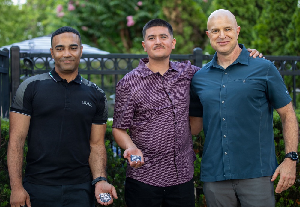 Three men are posing together outdoors in what appears to be a casual group photo. The man in the center is holding two small items that appear to be Army National Guard coins or badges. The setting features greenery, trees, and decorative iron fencing in the background.