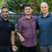 Three men are posing together outdoors in what appears to be a casual group photo. The man in the center is holding two small items that appear to be Army National Guard coins or badges. The setting features greenery, trees, and decorative iron fencing in the background.