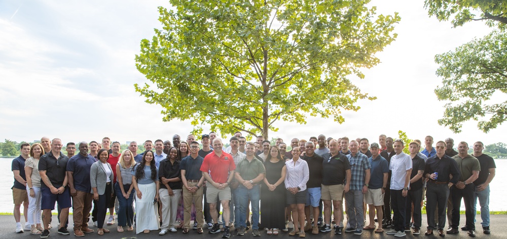 A large group of approximately 60-70 people are posed together for a formal group photograph outdoors near a body of water. The group appears diverse in age and gender, dressed in casual to business-casual attire, suggesting a professional or organizational gathering such as a company outing, reunion, or team event. The setting features a prominent large tree in the background with sunlight filtering through its leaves.