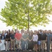 A large group of approximately 60-70 people are posed together for a formal group photograph outdoors near a body of water. The group appears diverse in age and gender, dressed in casual to business-casual attire, suggesting a professional or organizational gathering such as a company outing, reunion, or team event. The setting features a prominent large tree in the background with sunlight filtering through its leaves.