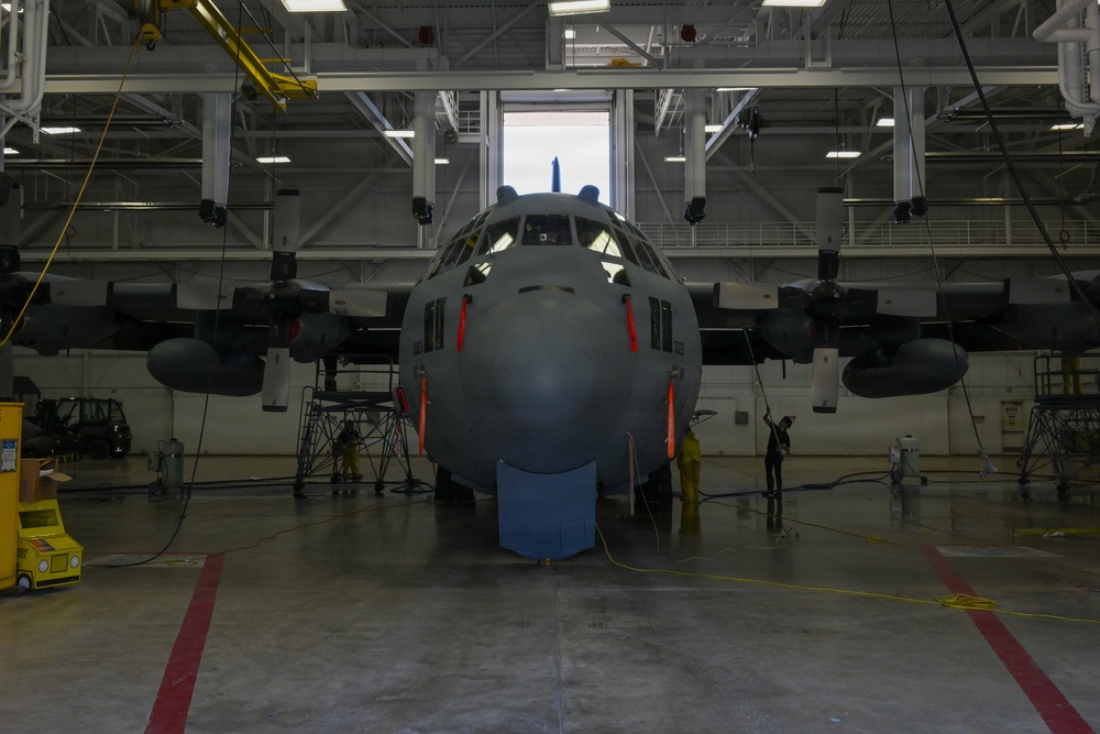 A large military turboprop aircraft, consistent with a C-130 Hercules variant, is positioned inside a large maintenance hangar undergoing washing or cleaning operations. Workers in yellow protective gear are visible on both sides of the aircraft's nose section, using hoses and equipment. The hangar is well-equipped with overhead cranes, lighting, and support infrastructure.