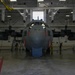 A large military turboprop aircraft, consistent with a C-130 Hercules variant, is positioned inside a large maintenance hangar undergoing washing or cleaning operations. Workers in yellow protective gear are visible on both sides of the aircraft's nose section, using hoses and equipment. The hangar is well-equipped with overhead cranes, lighting, and support infrastructure.