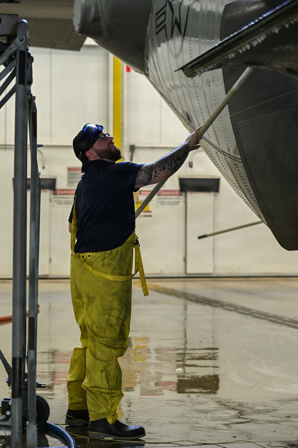 A male technician in yellow waterproof overalls is washing or scrubbing the underside of a large military or commercial aircraft inside a hangar facility. He is using a long-handled brush and looking upward at the aircraft surface, which shows visible rivets and panel lines. The wet floor and his protective gear indicate an active aircraft washing operation.