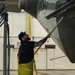 A male technician in yellow waterproof overalls is washing or scrubbing the underside of a large military or commercial aircraft inside a hangar facility. He is using a long-handled brush and looking upward at the aircraft surface, which shows visible rivets and panel lines. The wet floor and his protective gear indicate an active aircraft washing operation.