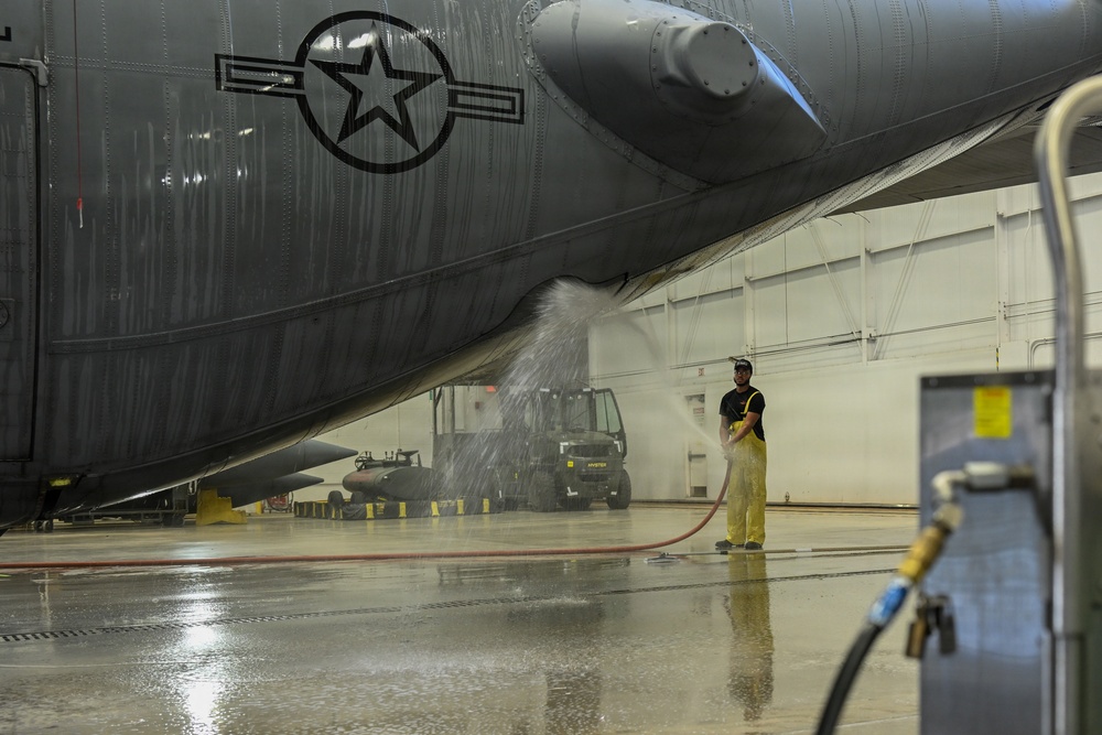 A maintenance technician in yellow protective overalls is washing the underside of a large military aircraft inside a hangar facility. The aircraft displays a US military star insignia on its fuselage and appears to be a large bomber or transport aircraft. The worker is using a red hose to spray water across the aircraft's belly in what appears to be a routine wash-down procedure.