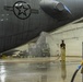 A maintenance technician in yellow protective overalls is washing the underside of a large military aircraft inside a hangar facility. The aircraft displays a US military star insignia on its fuselage and appears to be a large bomber or transport aircraft. The worker is using a red hose to spray water across the aircraft's belly in what appears to be a routine wash-down procedure.