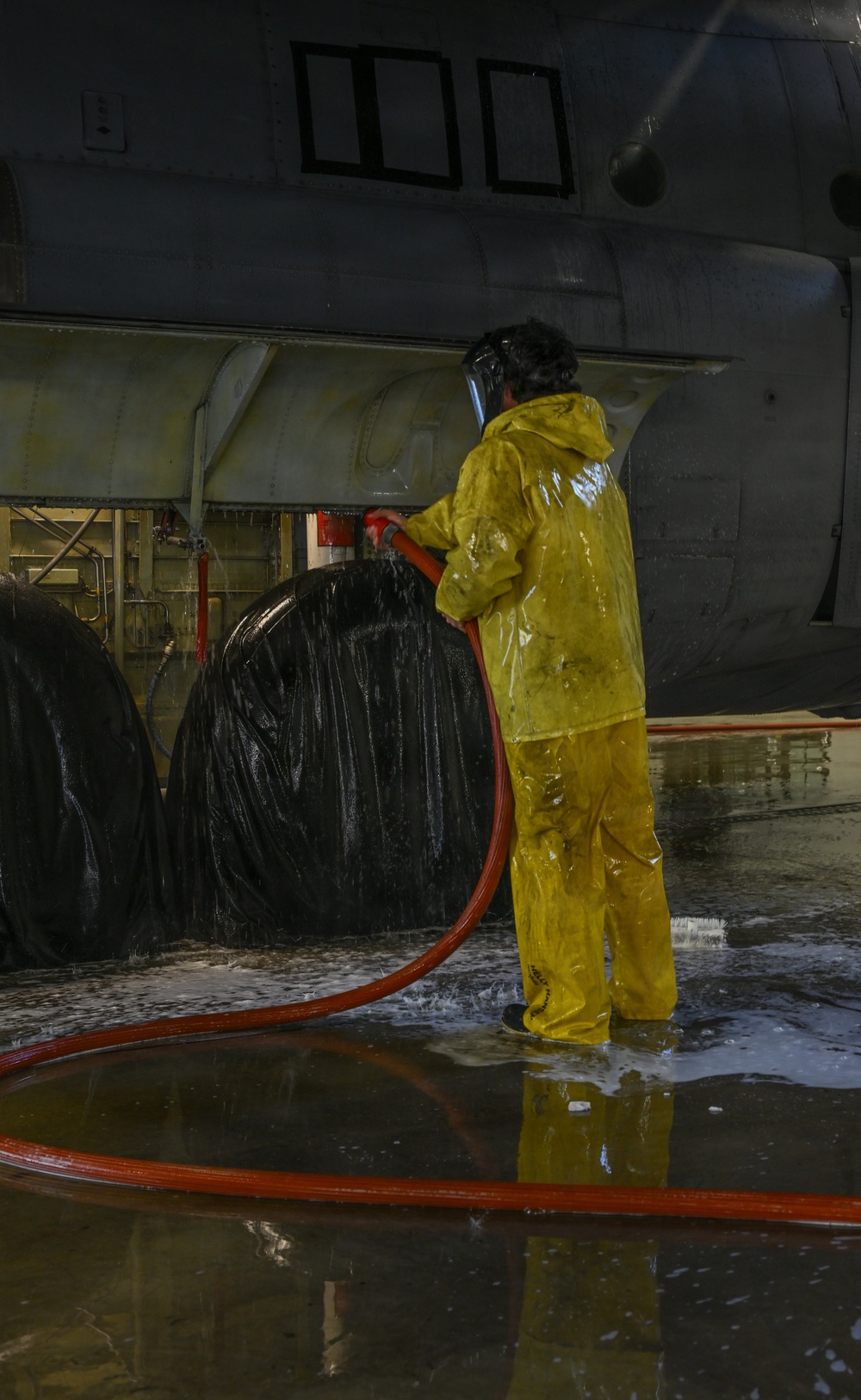 A worker in full yellow protective rain gear and a face shield is using a red hose to wash down large black rubber tires or fenders aboard what appears to be a military or naval vessel. The floor is flooded with water and soapy foam, indicating an active washdown operation. The setting appears to be an enclosed hangar deck or vehicle bay of a large ship.