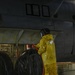 A worker in full yellow protective rain gear and a face shield is using a red hose to wash down large black rubber tires or fenders aboard what appears to be a military or naval vessel. The floor is flooded with water and soapy foam, indicating an active washdown operation. The setting appears to be an enclosed hangar deck or vehicle bay of a large ship.