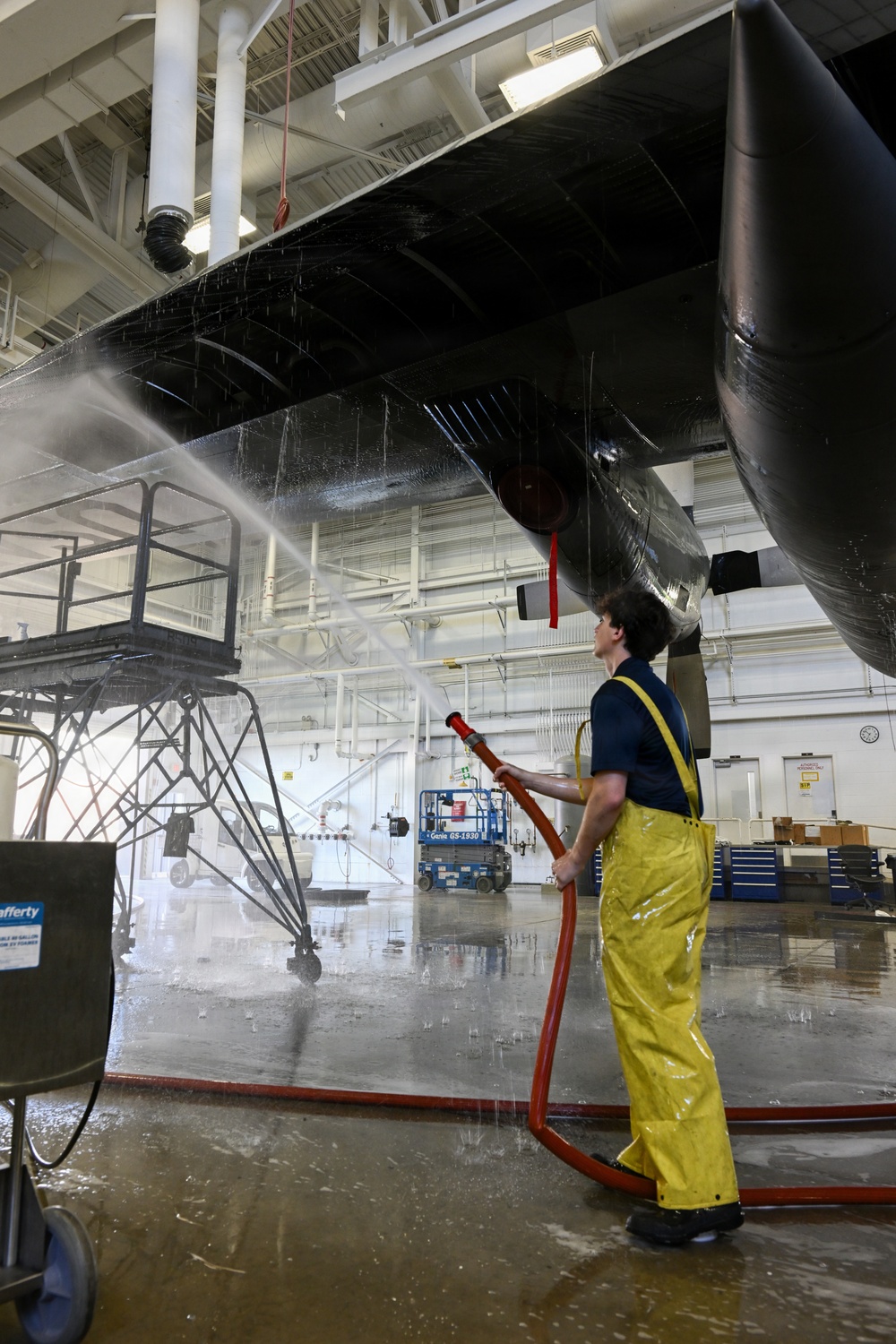 A worker in yellow waterproof overalls is washing the underside of a large black aircraft inside an indoor maintenance hangar. The worker is using a red hose to spray water at high pressure onto the aircraft's fuselage or wing underside. The facility appears to be a professional aviation maintenance or restoration facility with scissor lifts and work platforms visible in the background.