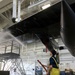 A worker in yellow waterproof overalls is washing the underside of a large black aircraft inside an indoor maintenance hangar. The worker is using a red hose to spray water at high pressure onto the aircraft's fuselage or wing underside. The facility appears to be a professional aviation maintenance or restoration facility with scissor lifts and work platforms visible in the background.