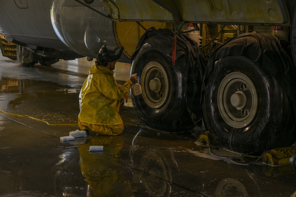 A maintenance worker in a yellow protective rain suit is kneeling on a wet hangar floor, cleaning the landing gear tires of a large aircraft. The worker appears to be applying a cleaning solution from a spray bottle to the massive multi-wheel landing gear assembly. The scene takes place indoors in what appears to be an aircraft maintenance hangar, with water and soap visible on the reflective floor.