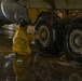 A maintenance worker in a yellow protective rain suit is kneeling on a wet hangar floor, cleaning the landing gear tires of a large aircraft. The worker appears to be applying a cleaning solution from a spray bottle to the massive multi-wheel landing gear assembly. The scene takes place indoors in what appears to be an aircraft maintenance hangar, with water and soap visible on the reflective floor.