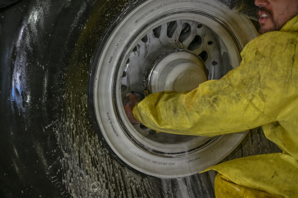 A person wearing a yellow protective suit is working on a large aircraft wheel and tire assembly. The wheel appears wet, suggesting cleaning or maintenance is being performed. The tire sidewall contains visible markings including size specifications and overhaul date information.