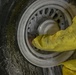 A person wearing a yellow protective suit is working on a large aircraft wheel and tire assembly. The wheel appears wet, suggesting cleaning or maintenance is being performed. The tire sidewall contains visible markings including size specifications and overhaul date information.