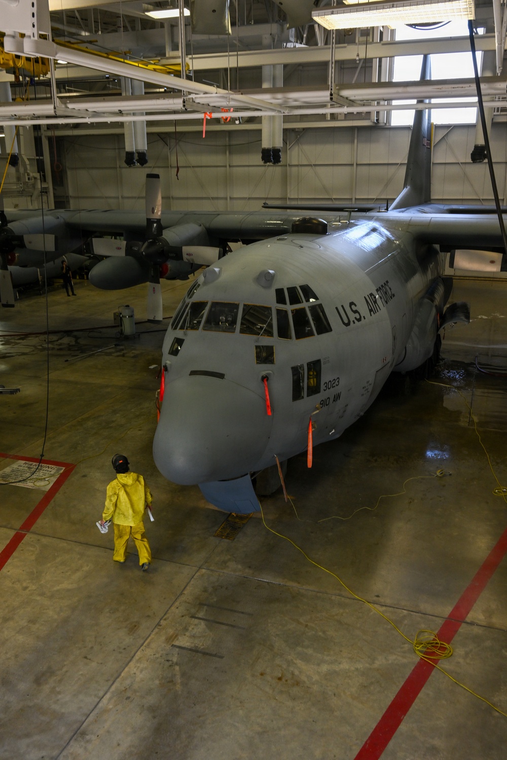 A U.S. Air Force C-130 Hercules transport aircraft (serial number 3023, assigned to the 910th Airlift Wing) is positioned inside a large maintenance hangar. A worker dressed in a yellow protective suit and helmet is standing near the nose of the aircraft, apparently conducting a wash or maintenance procedure. Yellow hoses and safety markings are visible on the hangar floor.