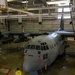 A U.S. Air Force C-130 Hercules transport aircraft (serial number 3023, assigned to the 910th Airlift Wing) is positioned inside a large maintenance hangar. A worker dressed in a yellow protective suit and helmet is standing near the nose of the aircraft, apparently conducting a wash or maintenance procedure. Yellow hoses and safety markings are visible on the hangar floor.